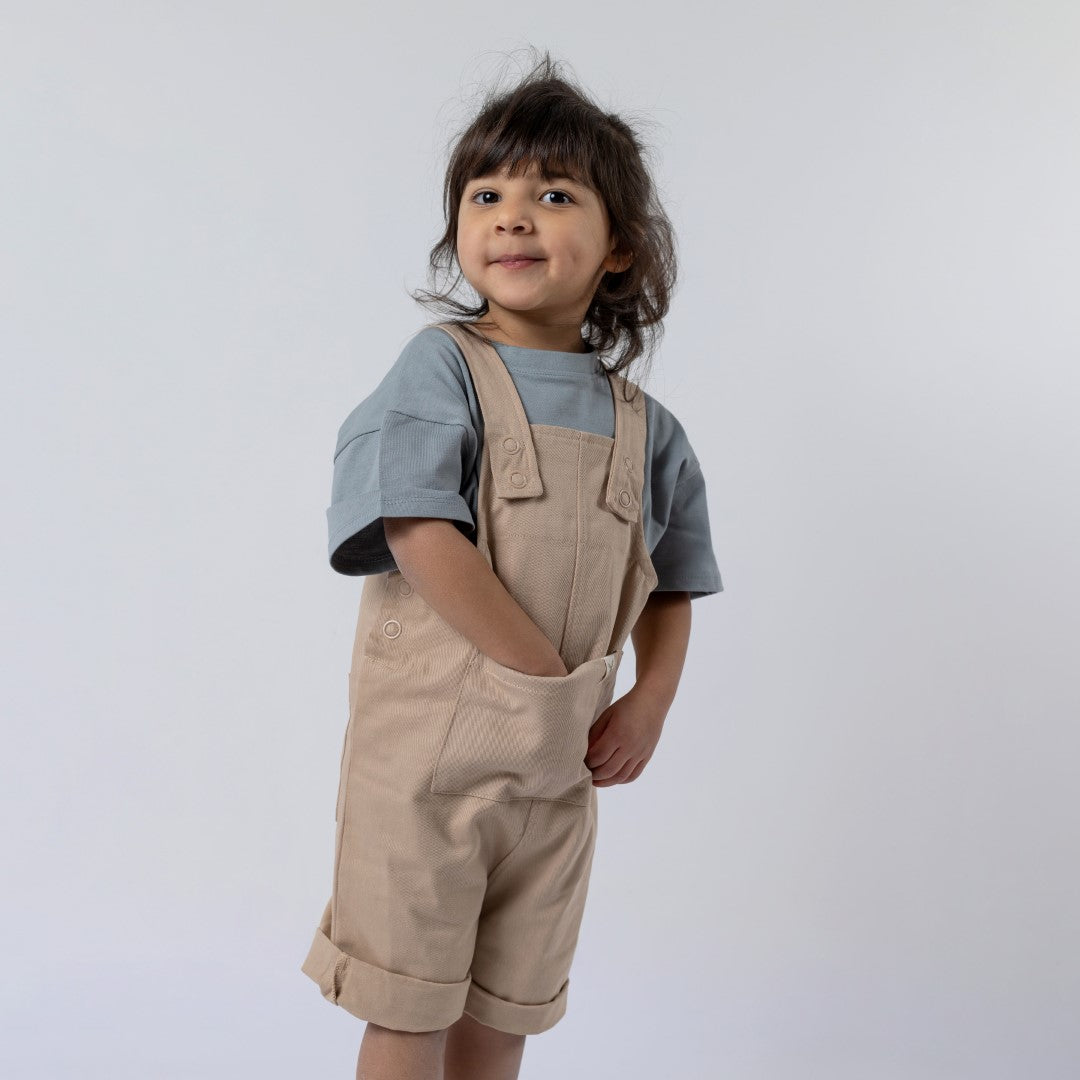 Young girl looking directly at the camera modelling beige dungaree shorts with hand in pocket