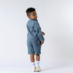 Small boy looking back at the camera, wearing a blue jacket with colour and matching shorts with the Aneby hem tag