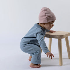 Small baby leaning against wooden stall whilst wearing blue Aneby jumpsuit and dusty pink beanie