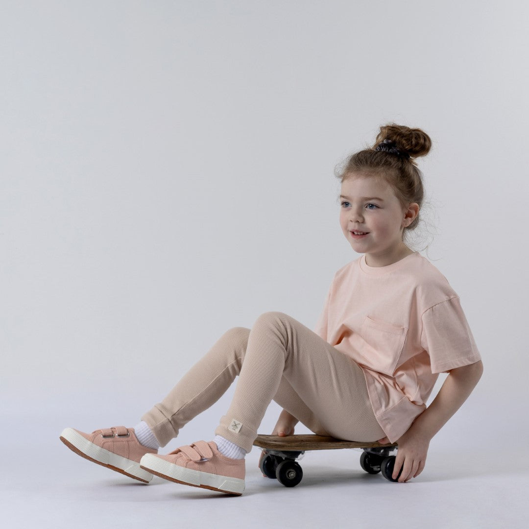 Girl sitting on skateboard looking to the side wearing beige leggings with rolled cuff and pink Aneby Tshirt