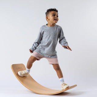 Young boy wearing long-sleeve tee with constellation design and pink shorts, balancing on a wooden wobble board