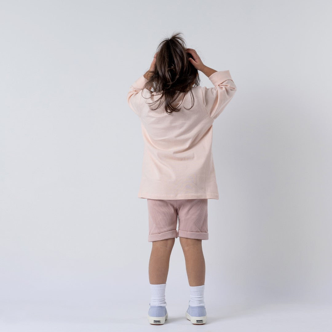 Young girl wearing pink long-sleeved t-shirt and shorts, with hands on her head and back to camera