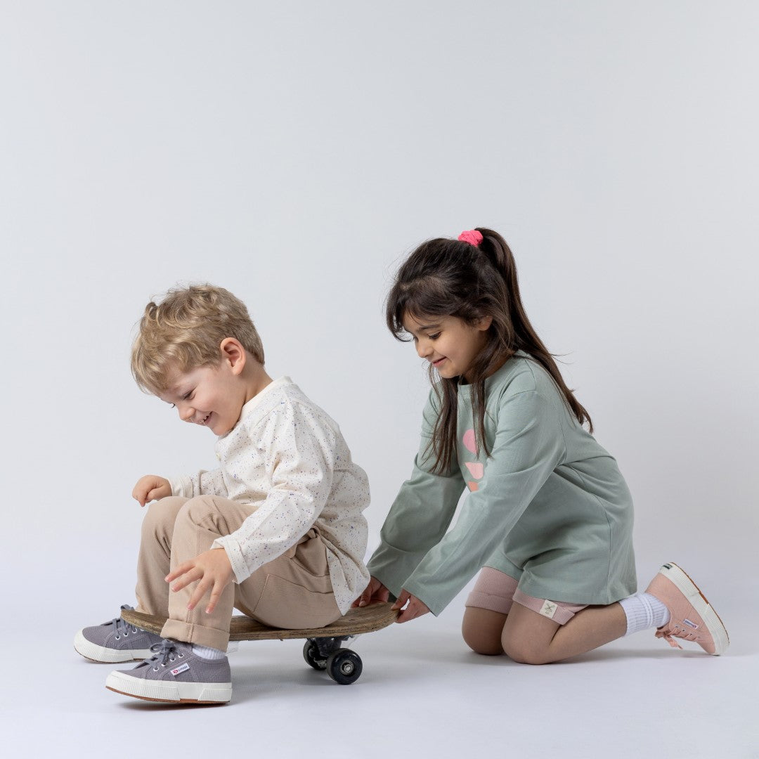 Young girl wearing teal long-sleeved t-shirt and pink shorts, standing behind small boy sitting on a skateboard