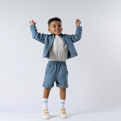 Boy with arms in the air wearing matching slate blue twill boxy jacket and shorts with speckled ecru tshirt