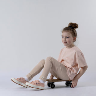 Young girl wearing a pink t-shirt featuring a front patch pocket sitting on a skateboard