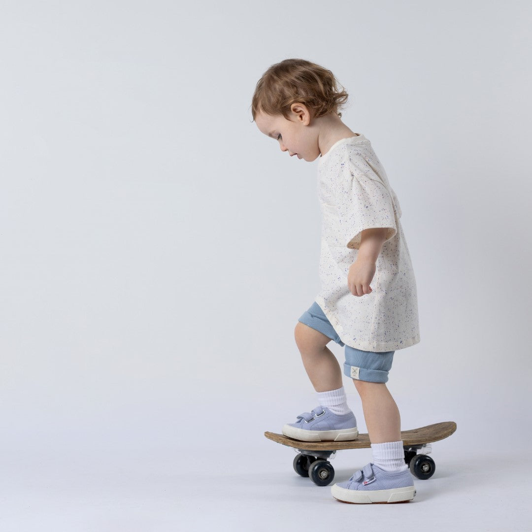 Small toddler trying to get her balance on a wooden skateboard whilst modelling the pastel speckled tee and blue chino shorts