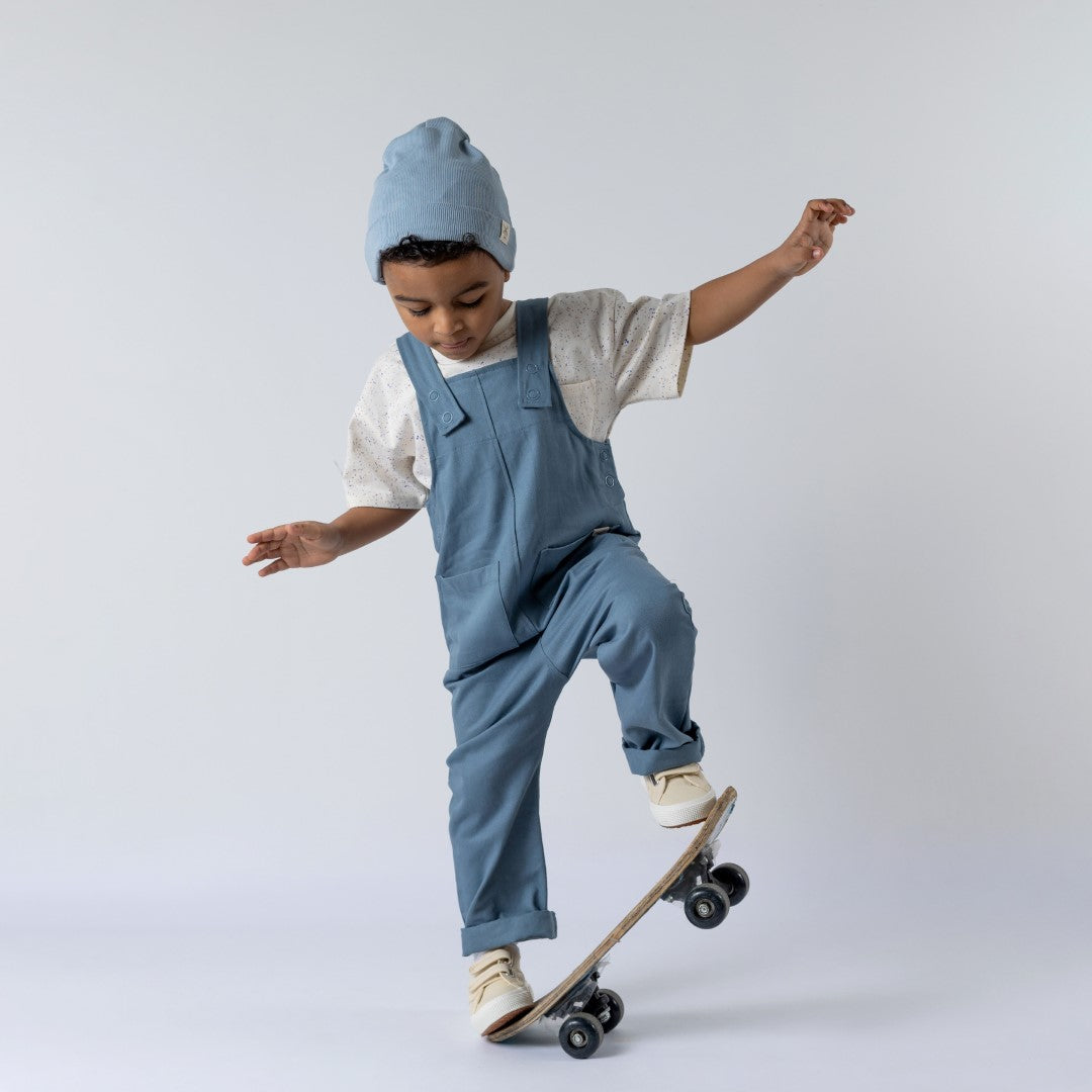 Young boy on a skateboard wearing the pastel speckled t-shirt beneath blue Aneby dungarees