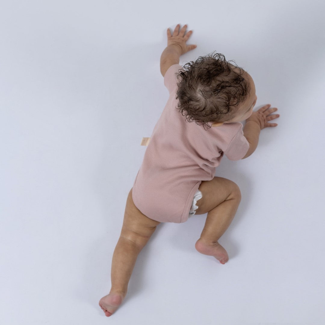 Bird’s eye view of a baby crawling across a neutral surface, wearing a dusty pink short sleeve baby bodysuit with ribbed texture.