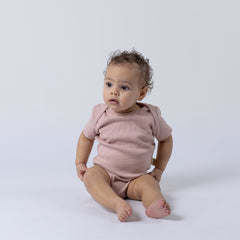 Small baby sitting upright and wearing a dusty pink short sleeve baby bodysuit, looking to the side against a softly lit, neutral background.