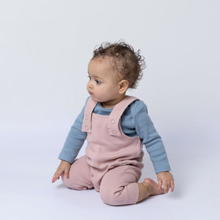 A baby wearing ribbed cotton dungarees in a dusty pink shade, shown from the front. The garment’s textured ribbing is clearly visible against a plain background.