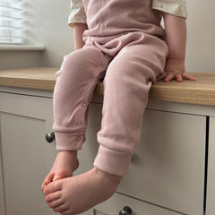 A toddler wearing ribbed cotton dungarees in dusty pink, shown from the front. The image captures the detailed ribbing and soft finish of the fabric, with the child seated on a wooden worktop in an indoor environment. 