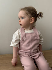 A girl wearing baby ribbed cotton dungarees and ecru t-short, sitting and looking to the side. She is seated on a wooden bench in a cosy indoor setting.