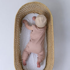 Small baby lying on their front in a moses basket, wearing a dusty pink sleeveless rib baby romper and a matching oat beanie hat. Neutral background.