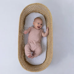 Small baby laughing while lying in a moses basket, wearing a dusty pink sleeveless rib baby romper. Neutral background.