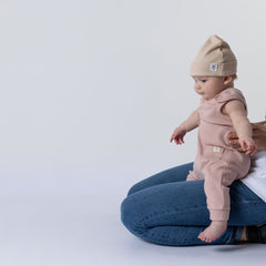 Small baby sitting on their mother’s knee, who is out of shot, wearing a dusty pink sleeveless rib baby romper and a matching oat beanie hat. Neutral background.