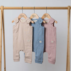 Three sleeveless rib baby rompers in dusty pink, slate blue, and oat hanging on a wooden frame. Neutral background.