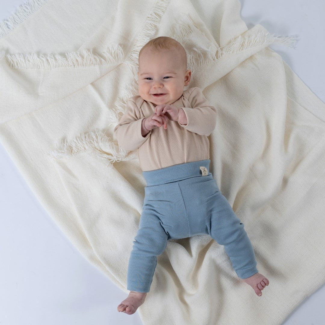 A smiling baby wearing organic ribbed baby leggings in slate blue, laying on a cream blanket, paired with an oat ribbed top. The baby is comfortable and relaxed, showing the soft texture of the leggings.