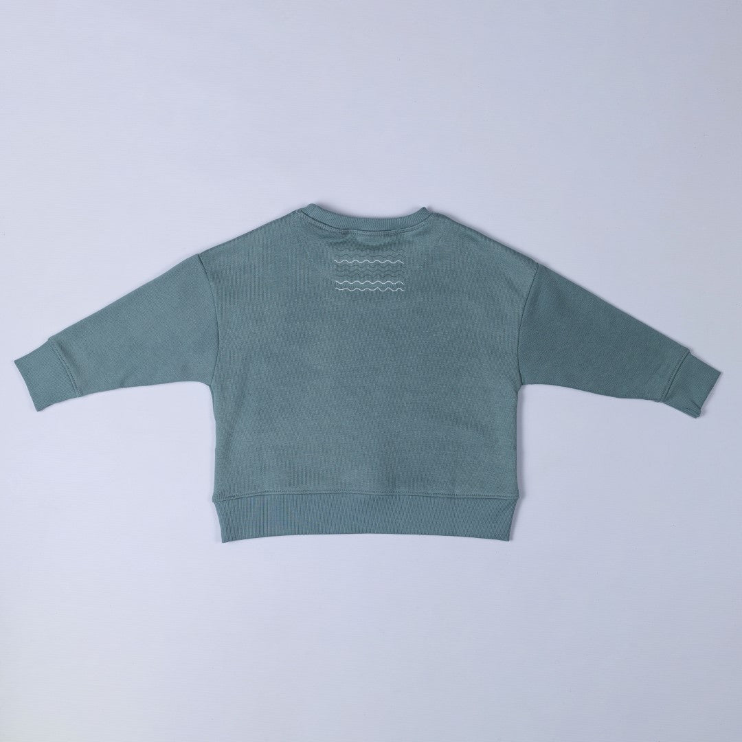 Flat lay of a steel blue sweatshirt, showing the back with a small white wave graphic below the collar, displayed against a neutral background.