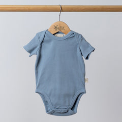Slate blue short sleeve baby bodysuit hanging neatly on a wooden hanger, made from ribbed organic cotton with soft rounded shoulders.