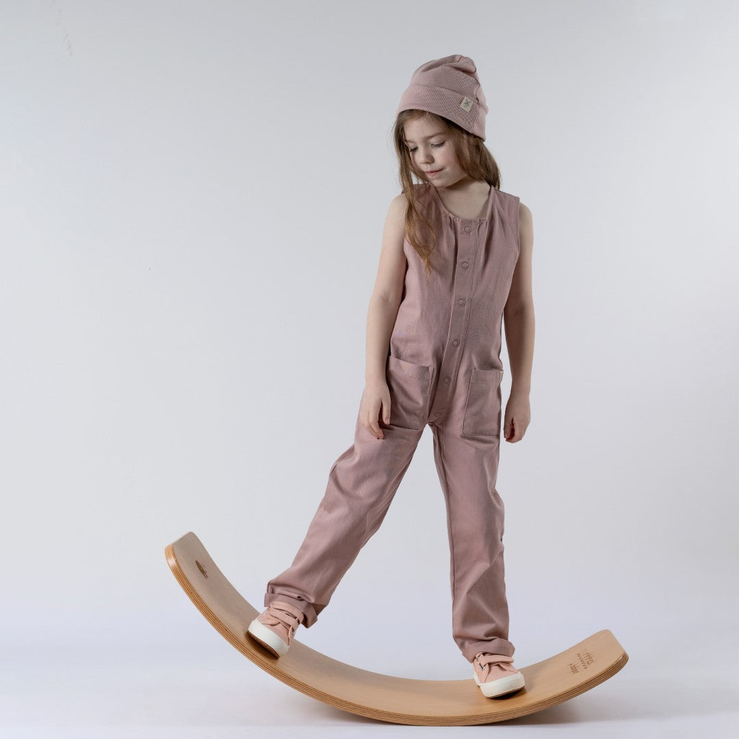 Young girl balancing on a wooden wobble board, wearing a dusty pink sleeveless rib baby romper and matching pink beanie hat. Hands by her side. Neutral background.