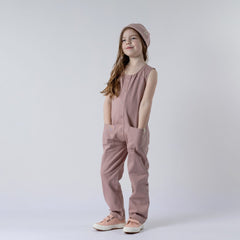 Young girl wearing a dusty pink sleeveless rib baby romper with hands in pockets, hair over shoulder, and matching pink beanie hat. Neutral background.