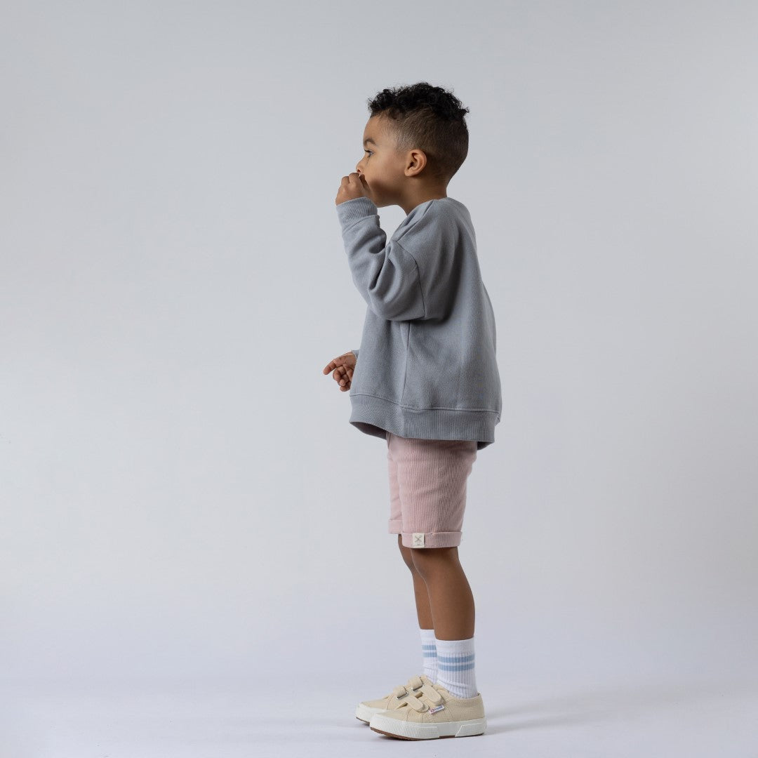 Young boy blowing bubbles, standing side-on against a neutral background, wearing dusty pink ribbed shorts with the Aneby hem tag visible and a grey jumper