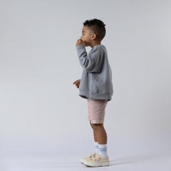 Young boy blowing bubbles, standing side-on against a neutral background, wearing dusty pink ribbed shorts with the Aneby hem tag visible and a grey jumper