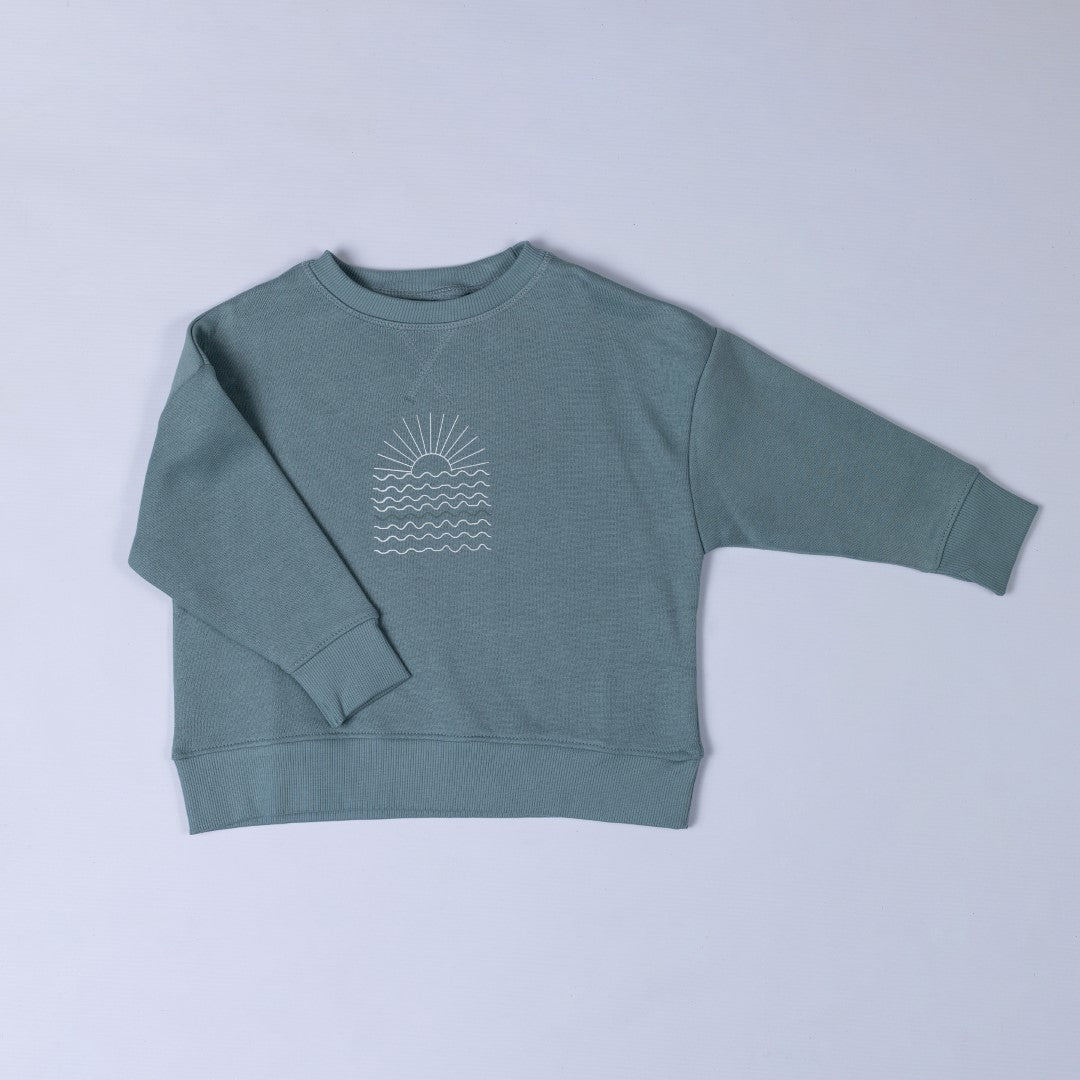 Flat lay of a steel blue sweatshirt featuring a white sunrise graphic on the front, displayed against a neutral background.