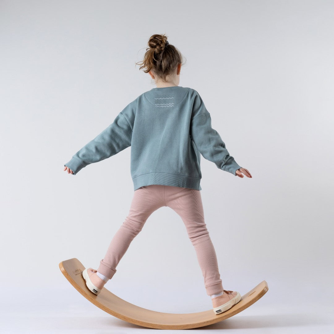 Young girl standing on a wooden wobble board, back to the camera, wearing a steel blue sweatshirt with a white wave graphic below the collar and pink leggings. Neutral background.