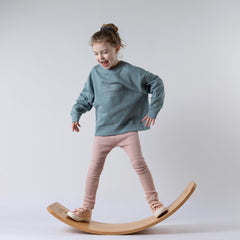 Young girl balancing on a wooden wobble board, smiling, wearing a steel blue sweatshirt with a white sunrise graphic on the front, paired with pink leggings. Neutral background.
