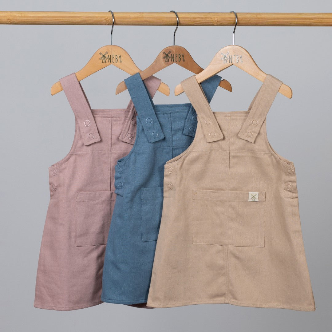 A set of three organic cotton dungaree dresses in dusty pink, slate blue, and oat, hanging from wooden clothes frame with Aneby clothes hangers