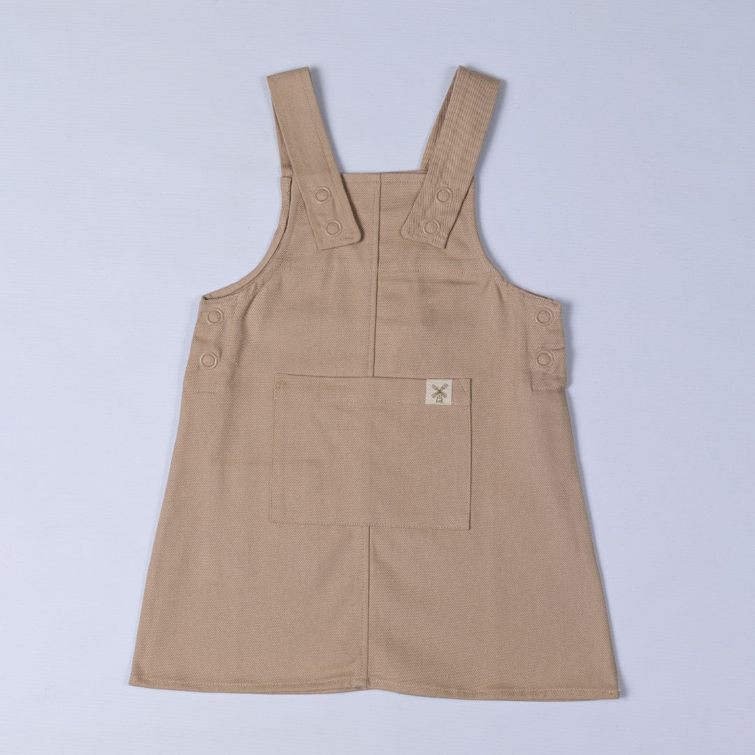 Flat lay of an oat-coloured organic cotton dungaree dress, highlighting the front pocket, sturdy straps, and soft twill fabric.