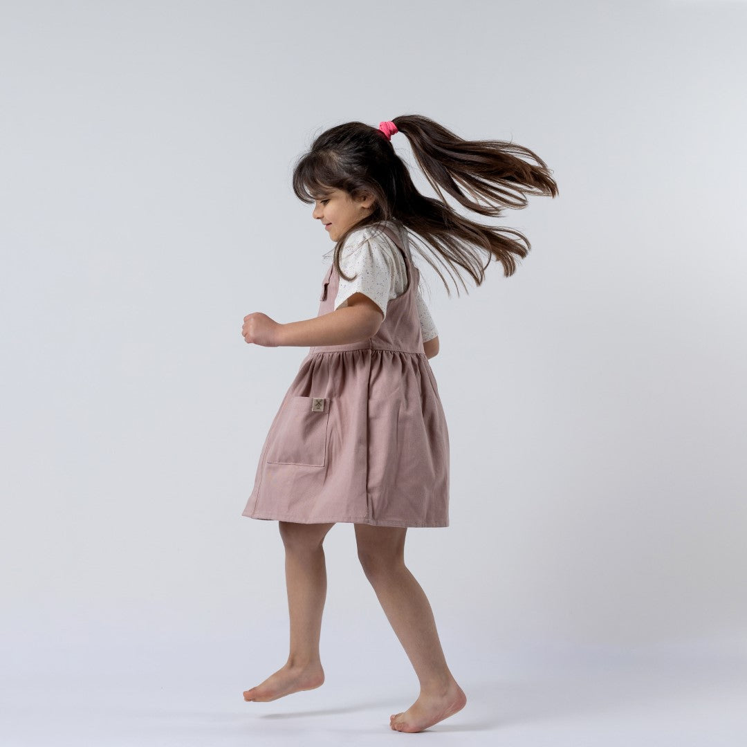 A dusty pink girls’ pleated dress with straps, displayed on a child. The dress features a flowing pleated skirt and delicate straps, perfect for special occasions.