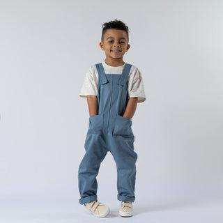 A boy wearing kids dungarees with patch pockets in blue twill. He is shown in a relaxed, natural pose against a plain white background. The image highlights the durable fabric, clear patch pocket details, and precise stitching, with natural lighting that accentuates the garment’s texture.