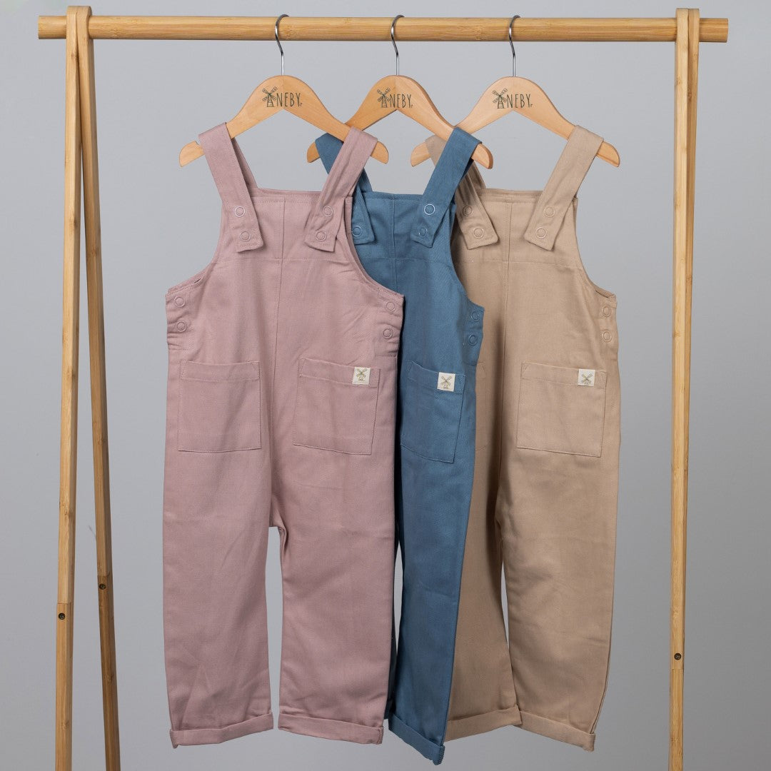 Product image displaying a set of three kids dungarees with patch pockets in blue, beige, and pink. The garments are neatly arranged on a wooden clothes rail on wooden hangers, highlighting their patch pocket details and fabric texture. 