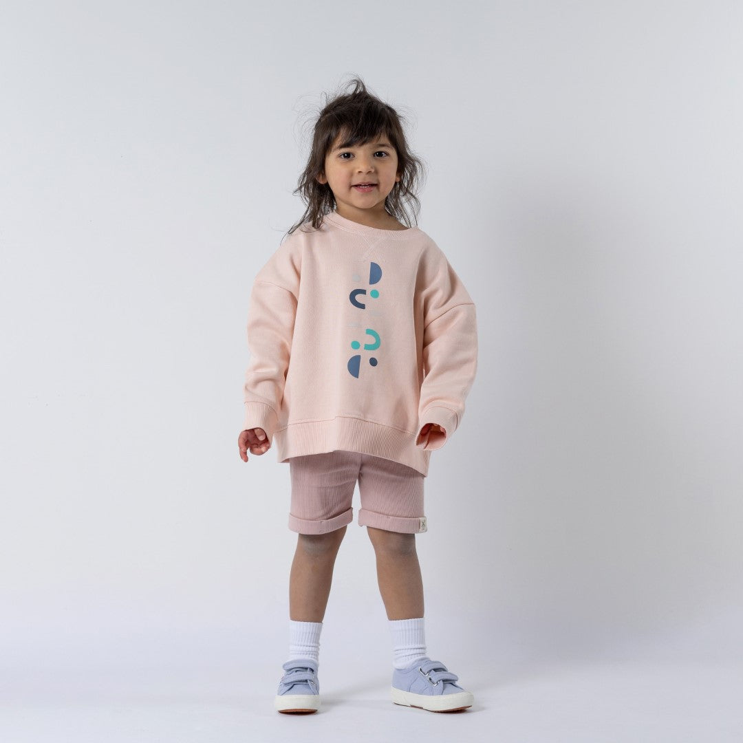 Young girl standing face-on, looking directly at the camera, wearing a kids pink sweatshirt with matching pink shorts. Neutral background.
