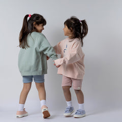 Two young sisters playing, holding hands and laughing. One wears a green sweatshirt with blue shorts, the other wears a kids pink sweatshirt with matching pink shorts. Neutral background.
