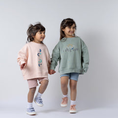 Two sisters holding hands and smiling. The younger girl wears a kids pink sweatshirt, with her outfit in focus. Neutral background.