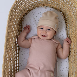 Smiling baby lying in a woven moses basket, wearing an oat kids ribbed cotton beanie hat and pink ribbed romper.