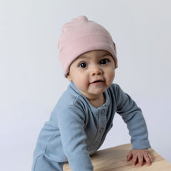 A baby wearing a blush pink kids ribbed cotton beanie hat and a slate blue ribbed cotton sleepsuit, leaning on a wooden surface against a light background.