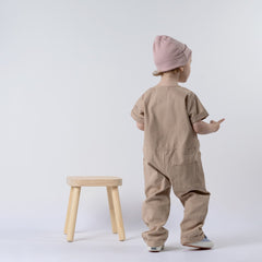 Small girl wearing an oat short-sleeve overall and pink beanie, with her back to the camera, standing in front of a wooden stool and neutral background.