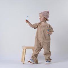 Small girl holding a bubble wand, wearing an oat short-sleeve overall with a pink beanie, standing in front of a wooden stool and neutral background.