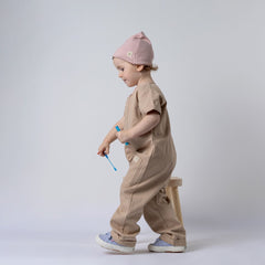 Side view of a small girl wearing an oat short-sleeve overall with a pink beanie, standing next to a wooden stool in a neutral background.