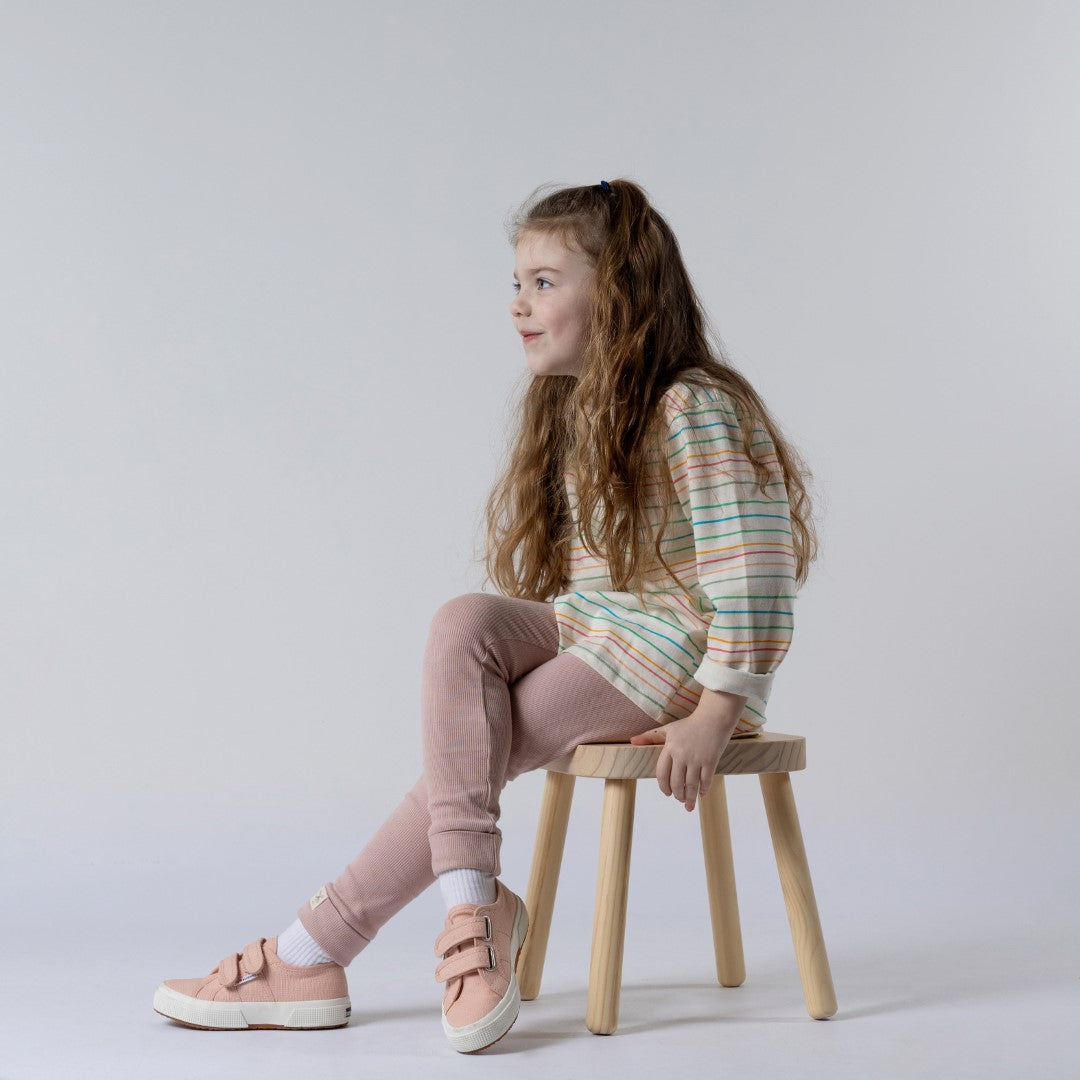 Young girl sitting cross-legged on a wooden stool wearing a kids long sleeve striped t-shirt with pastel blue, pink and yellow stripes, and dusty pink leggings.
