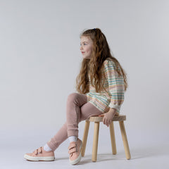 Young girl sitting cross-legged on a wooden stool wearing a kids long sleeve striped t-shirt with pastel blue, pink and yellow stripes, and dusty pink leggings.