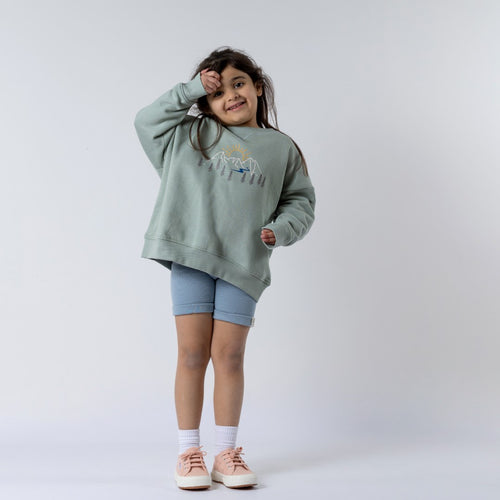 A child wearing a sage green kids' sweatshirt with a mountain print, standing against a neutral background. The sweatshirt has a relaxed fit with ribbed cuffs and hem.