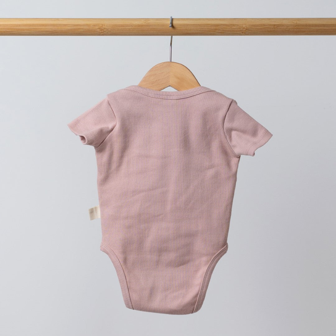 Dusty pink short sleeve baby bodysuit hanging from a wooden hanger, back view showing clean finishing and soft breathable cotton fabric.