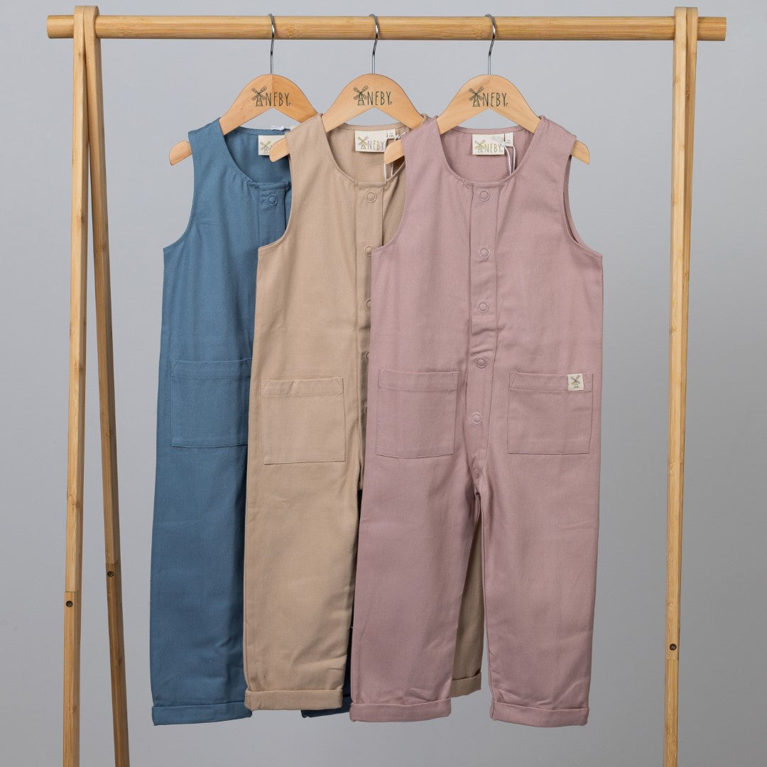 Three sleeveless rib baby rompers in dusty pink, slate blue, and oat, displayed on a wooden clothes frame against a neutral background.