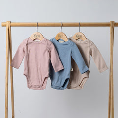 Three ribbed bodysuits with long sleeves for babies in oat, dusty pink, and slate blue, hanging neatly on wooden hangers on a wooden clothes rail.