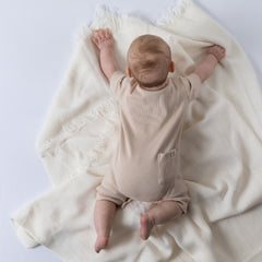 Baby lying on stomach, wearing an oat short sleeve rib baby romper with back pocket and Aneby hem tag visible, arms spread out wide.